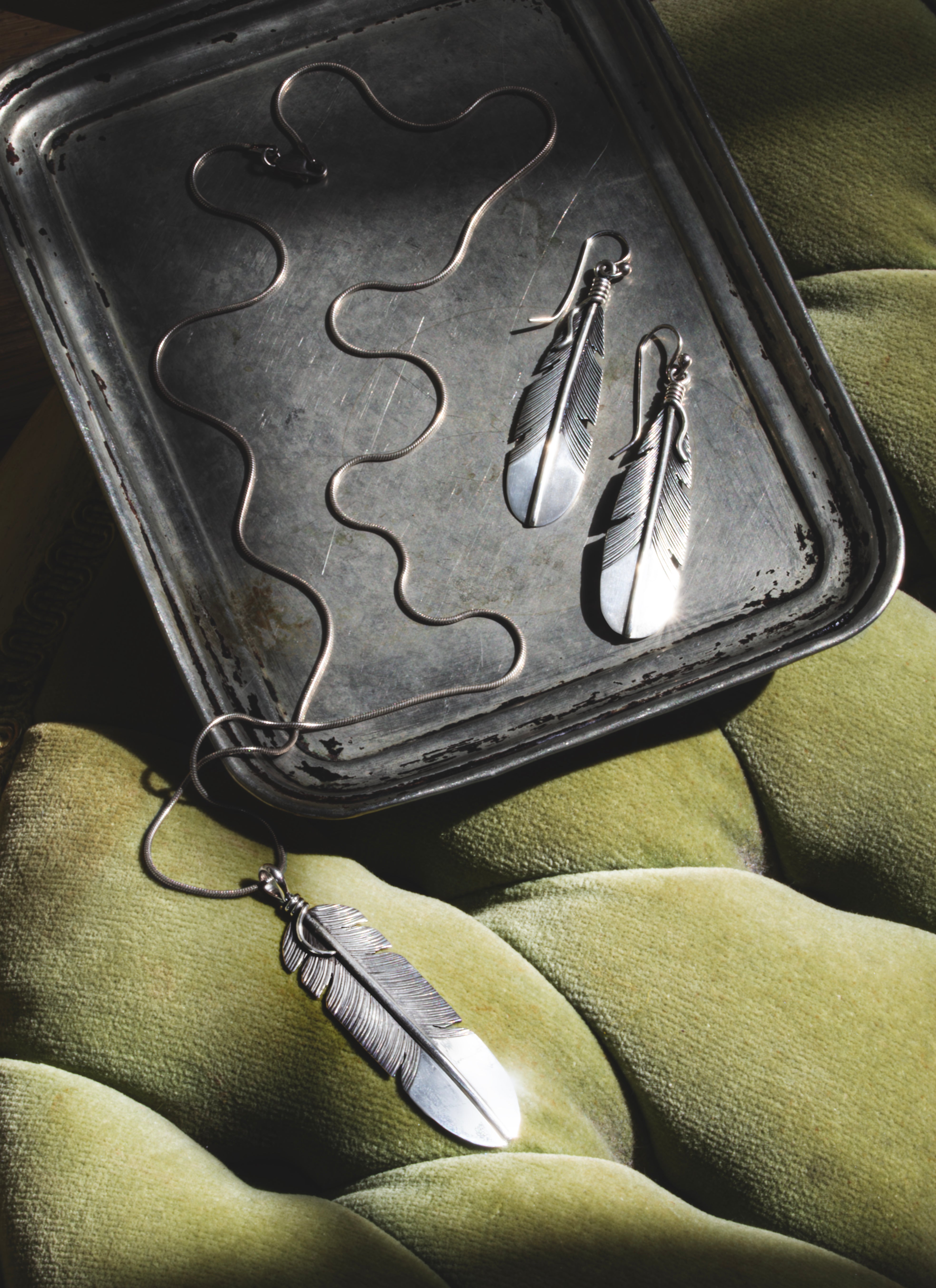 image of silver necklace and earrings shaped like feathers on a silver tray with a green plush background
